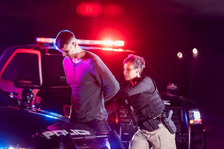 A police officer arrests a man, who has his hands behind his back, in front of a police car with flashing lights. The image illustrates a page section about unlawful searches.